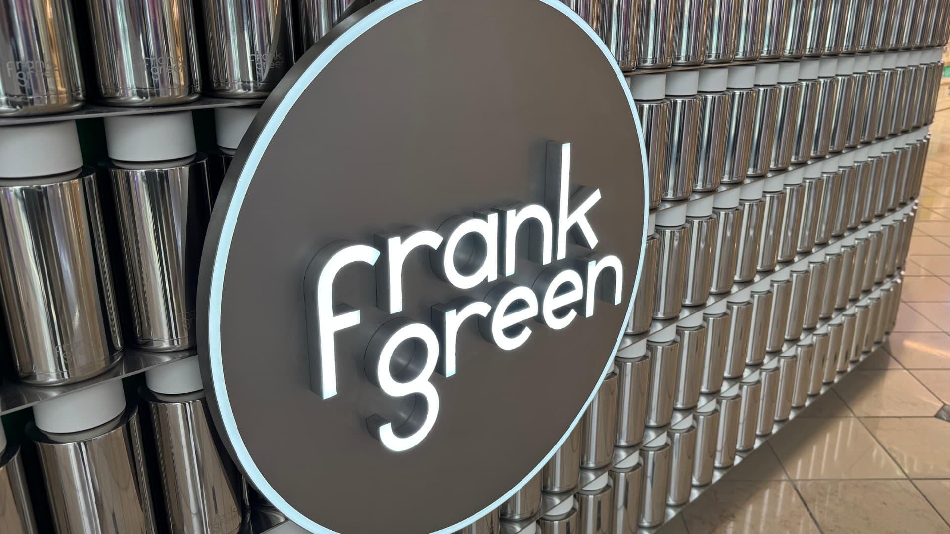 Illuminated circular Frank Green logo on a wall of silver water bottles