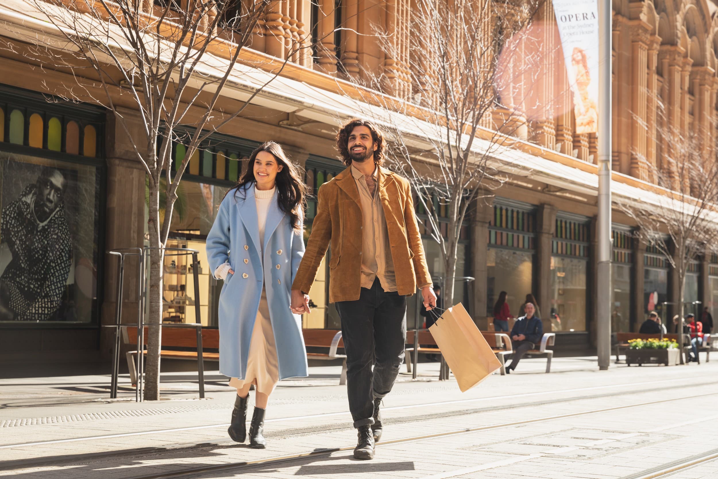 Two people walk away from a shopping centre hand in hand carrying a shopping bag